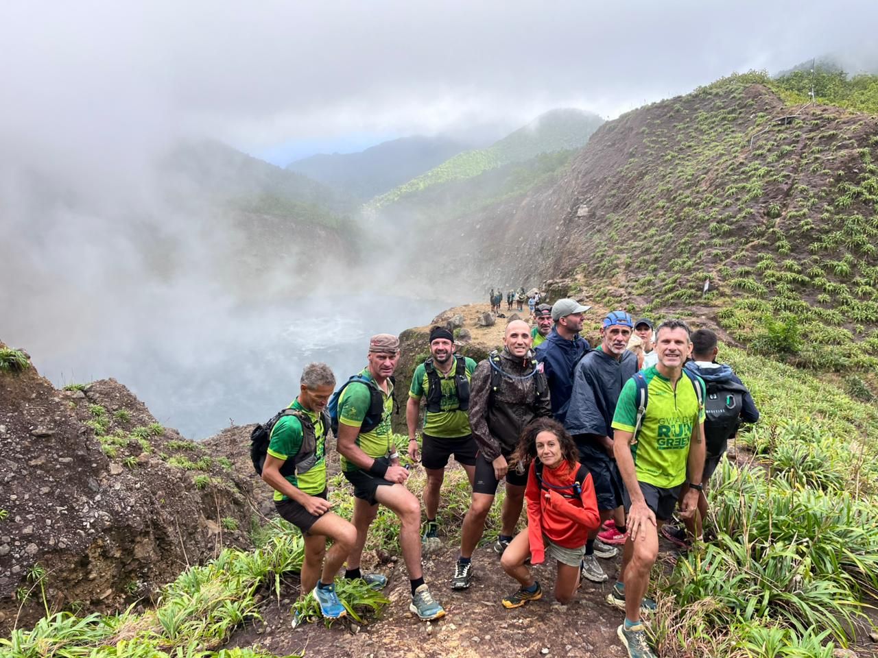 Les membres de la Team Gwada Run Green en maillot vert posent au bord d'un cratère fumant, entre brume et versants verdoyants